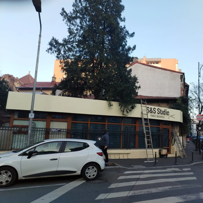 Montaj banner publicitar client salon barbershop pe atic fatada salon barbershop din Bucuresti — echipa Mora Group cu scara metalica profesionala, vedere exterioara la intersectia cu trecere pietoni si trafic auto local