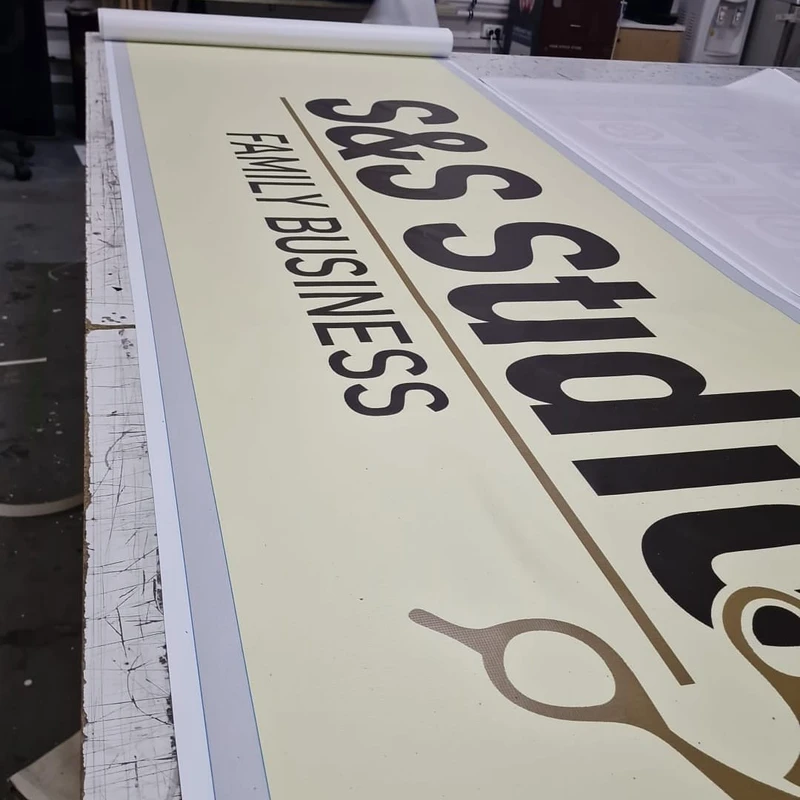 Detaliu close-up banner client salon barbershop pe masa atelier Mora Group — verificare calitate tipografie si foarfeca aurie, print eco-solvent 1440 dpi pe PVC frontlit fara halo sau granulatie vizibila