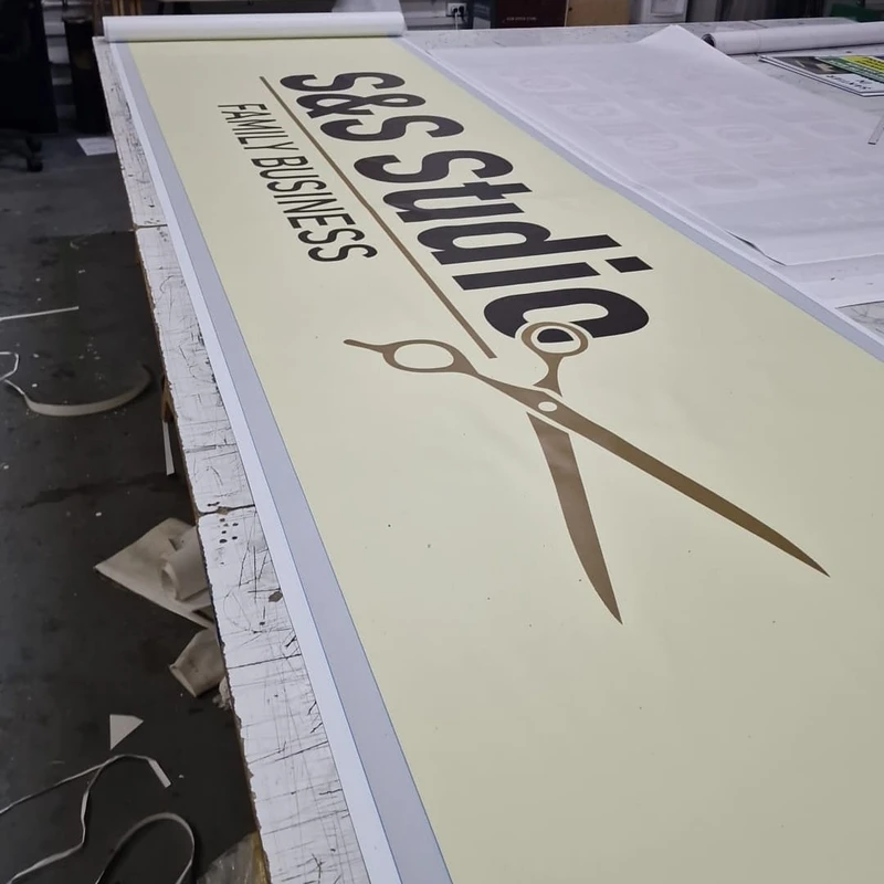 Banner client salon barbershop finalizat in atelierul Mora Group — print large-format pe PVC frontlit cu logo brand, foarfeca aurie si tipografie negru/gold, vedere oblica completa pe masa de productie inainte de pliere si livrare