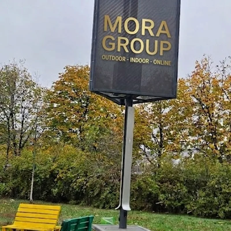 Detaliu close-up al stalpului solar Mora Group cu panou fotovoltaic in varf si banner publicitar pe negru cu accent gold, instalat la scoala gimnaziala