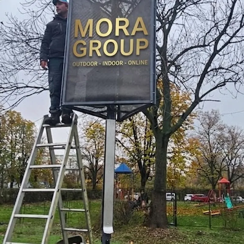 Echipa Mora Group monteaza stalpul solar cu banner publicitar in curtea scolii gimnaziale, folosind scara pentru fixarea panoului fotovoltaic si a casetei luminoase